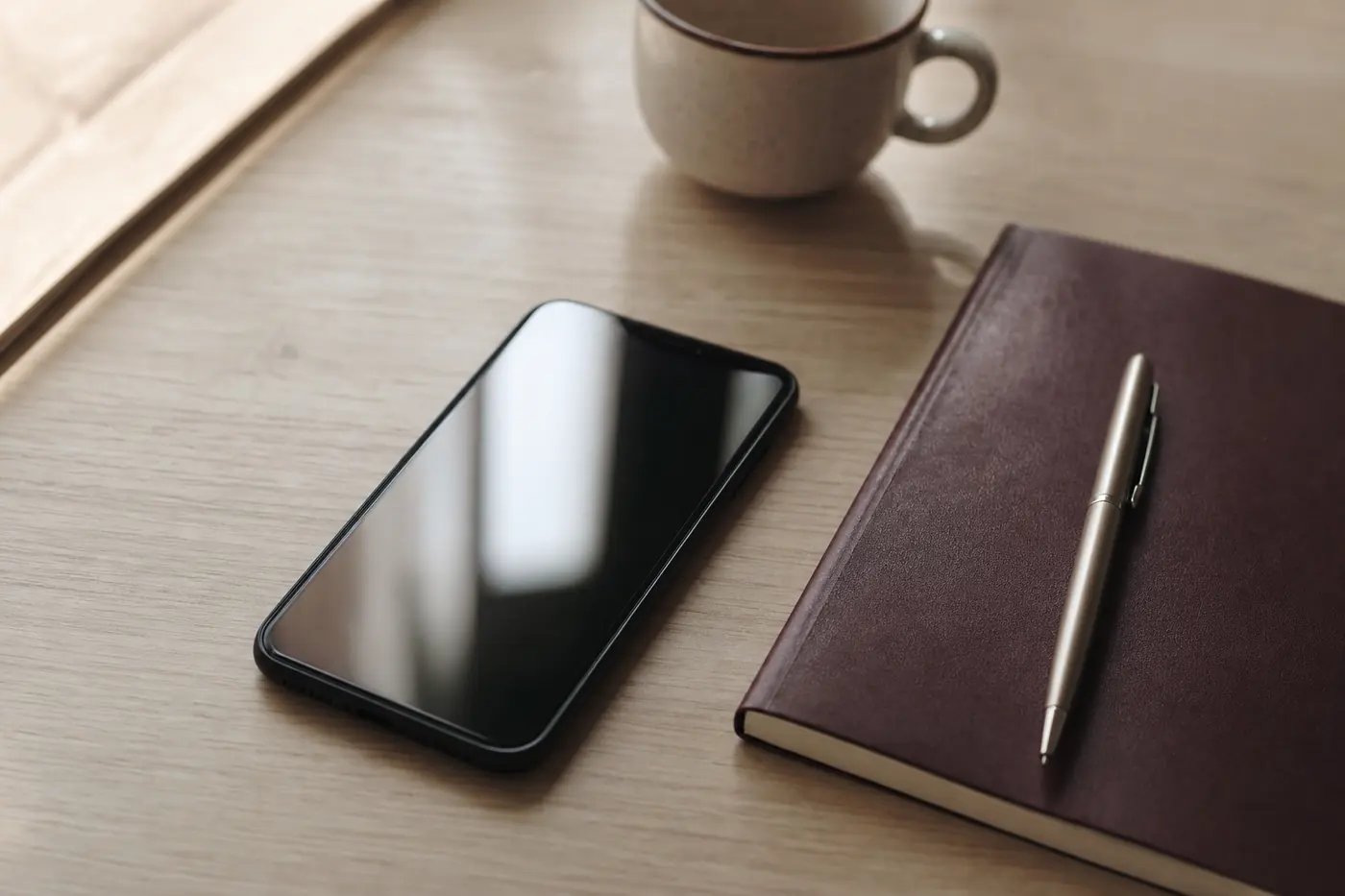 celular com a tela apagada sobre a mesa de madeira, ao lado de um caderno fechado e uma caneta